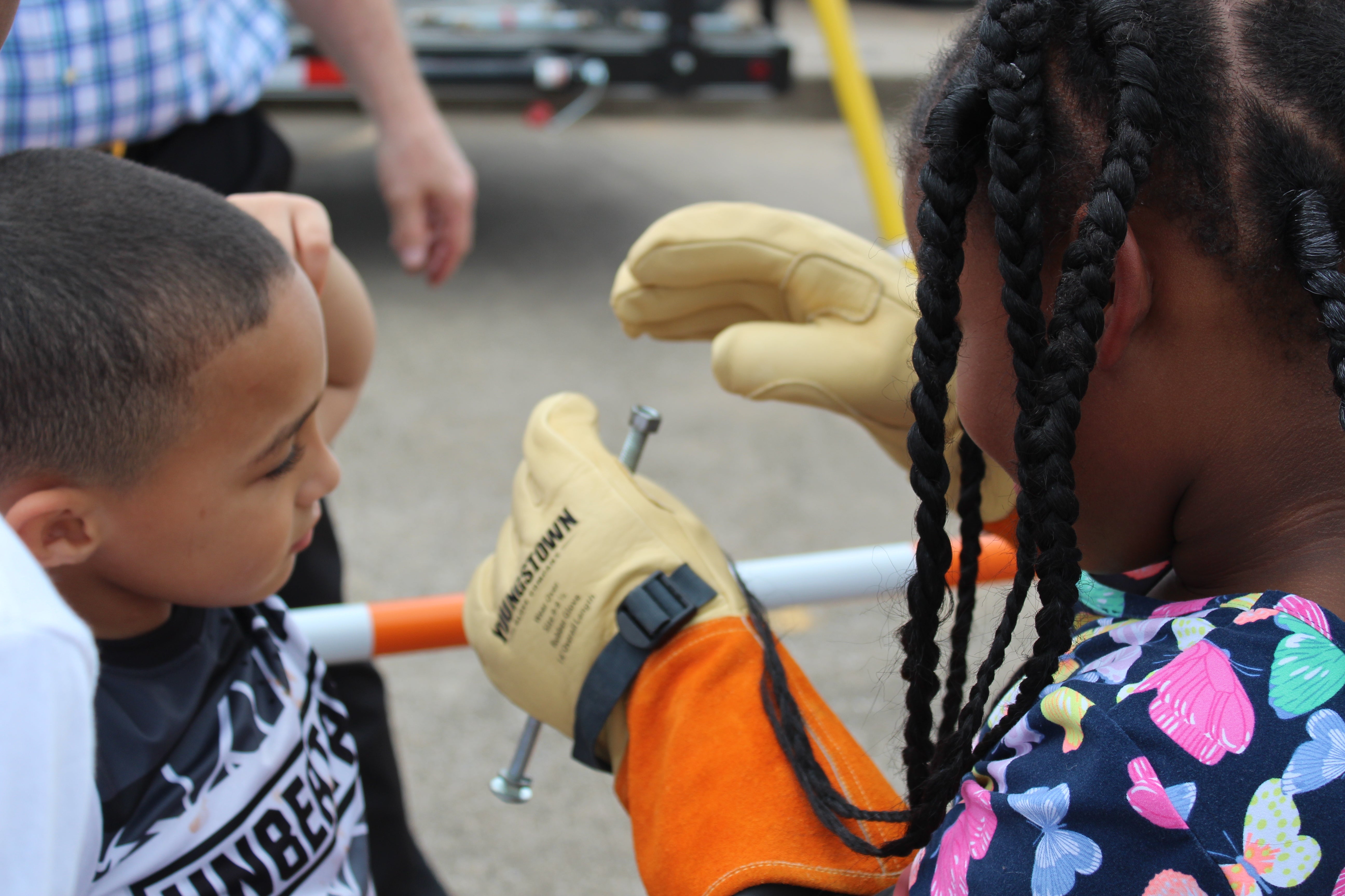 A young boy watches as a young girl attempts to put a nut on a bolt while wearing linemen gloves.